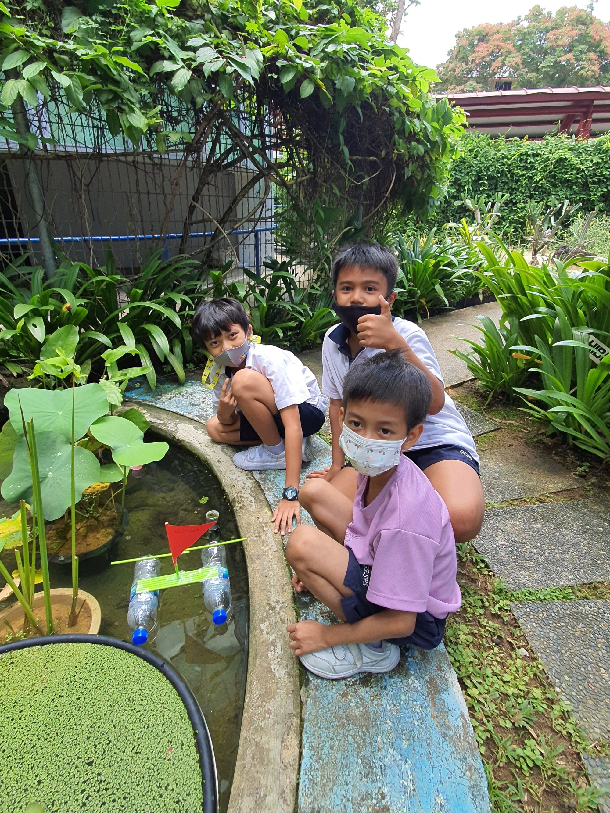 Three masked boys sit beside a garden pond with a small homemade bottle raft bearing a red flag.